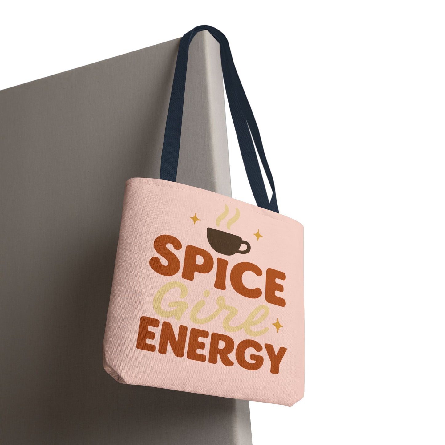 “Spice Girl Energy” tote bag in pale pink with navy straps hanging on wall — coffee-themed pastel tote for gifting and daily errands.