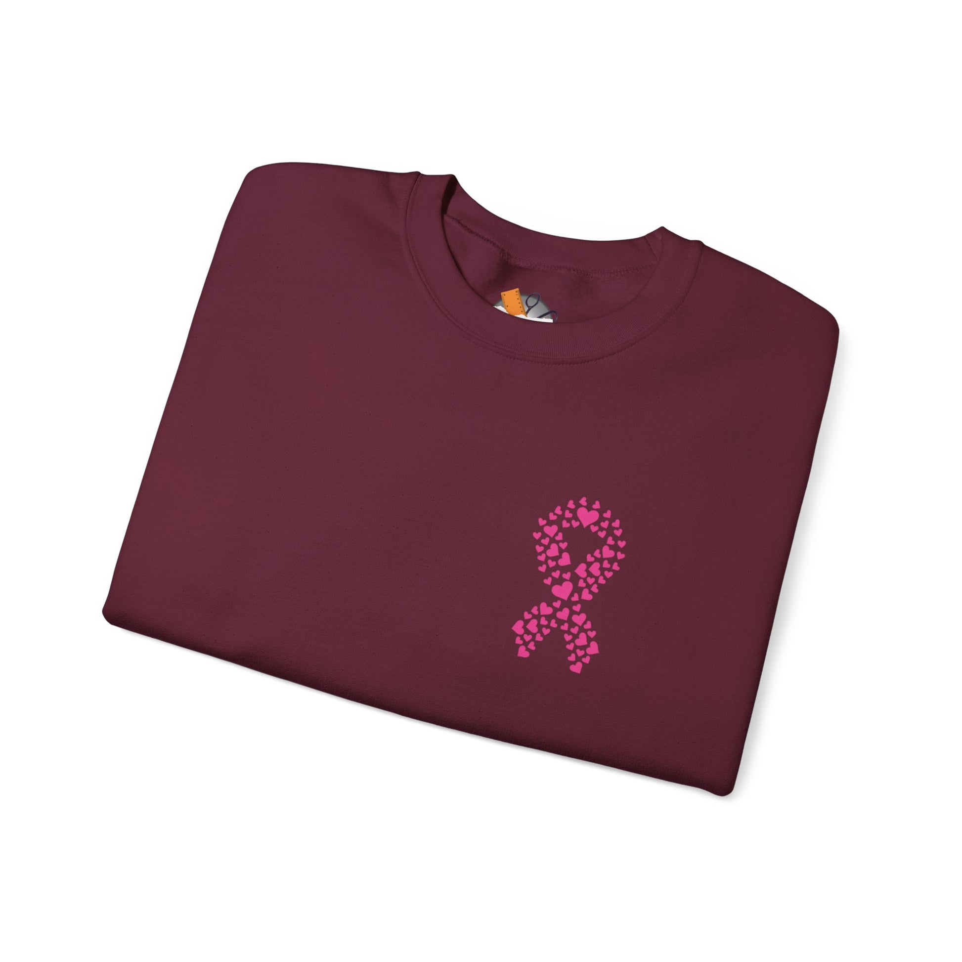 Folded maroon crewneck sweatshirt featuring pink heart-shaped ribbon design for breast cancer awareness, Makin’ It by Lady Ev.