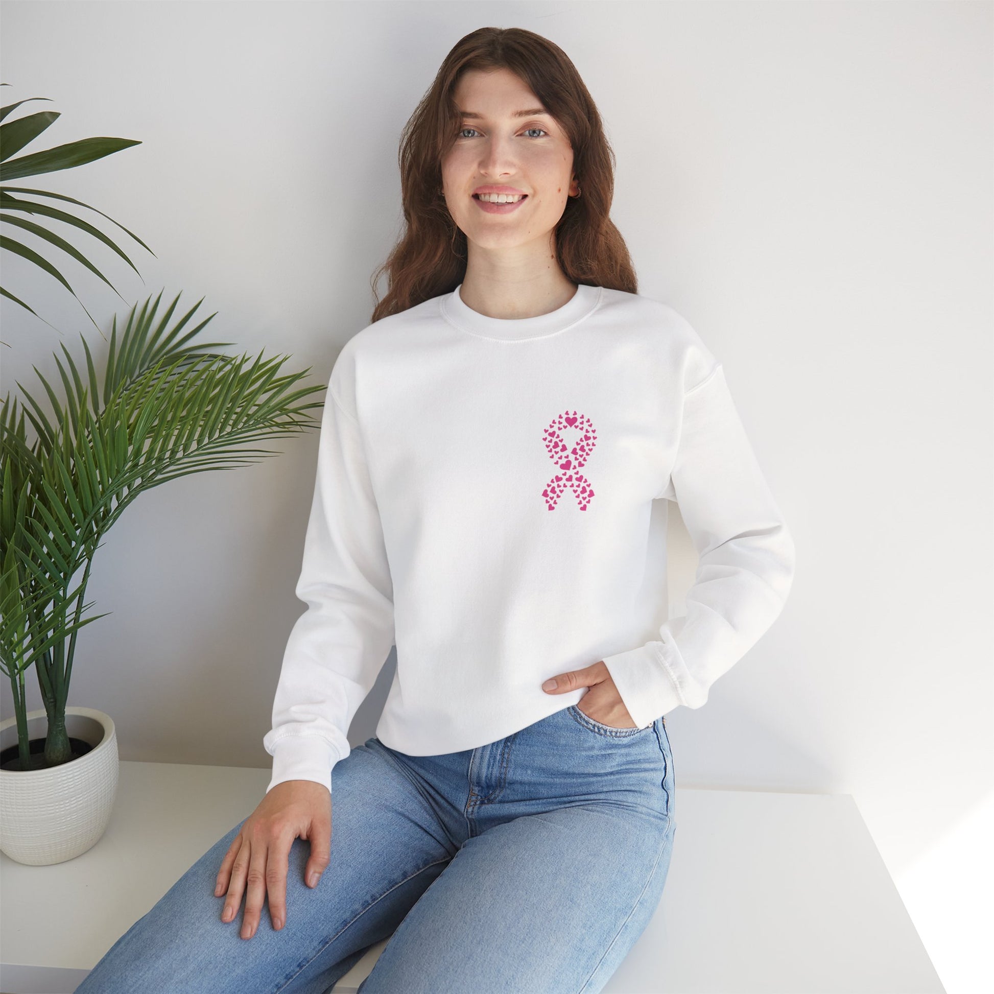 Woman seated in white breast cancer awareness crewneck sweatshirt featuring pink ribbon heart graphic, Makin’ It by Lady Ev.