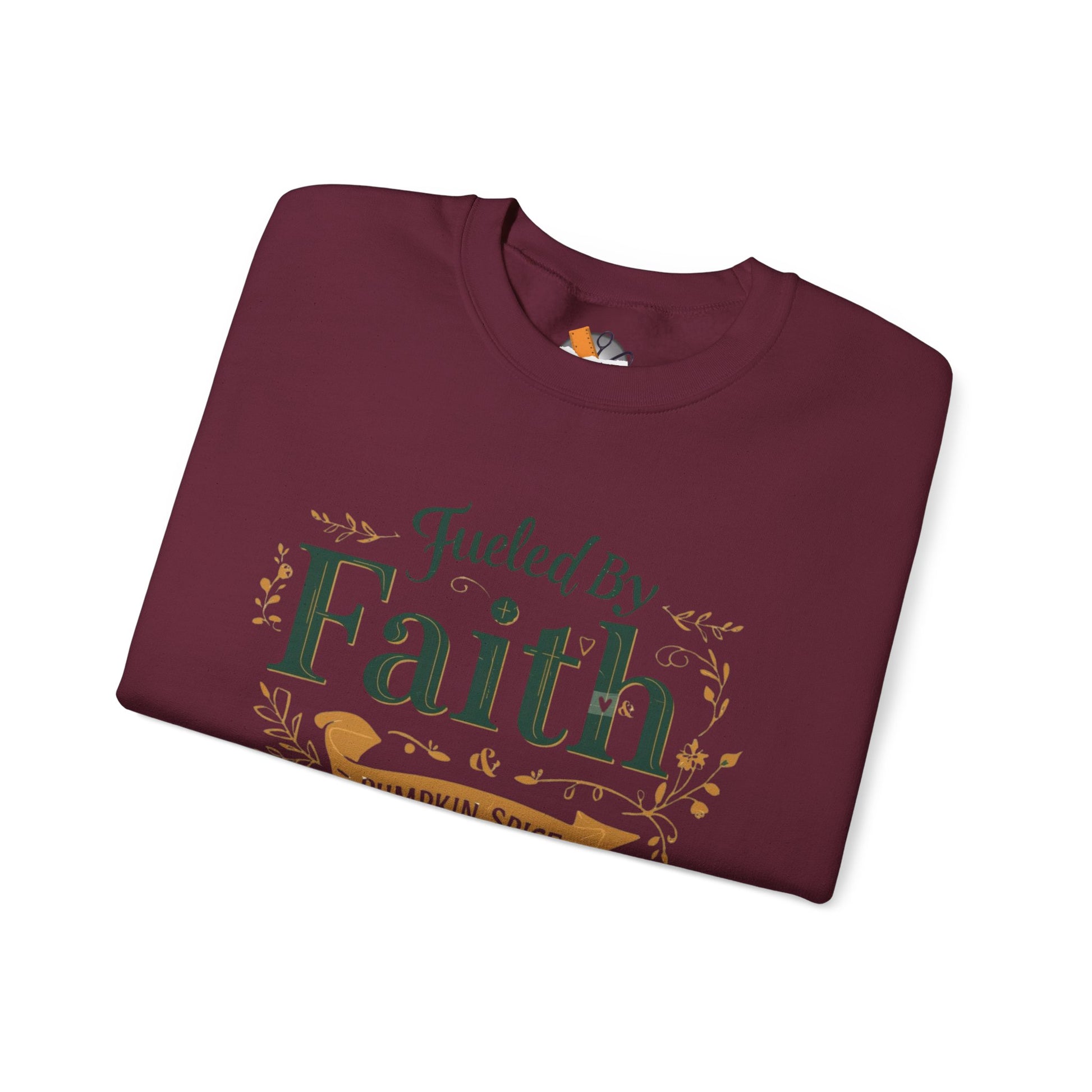 Folded maroon “Fueled by Faith & Pumpkin Spice” sweatshirt with gold and green design — cozy Christian fall apparel flat lay.