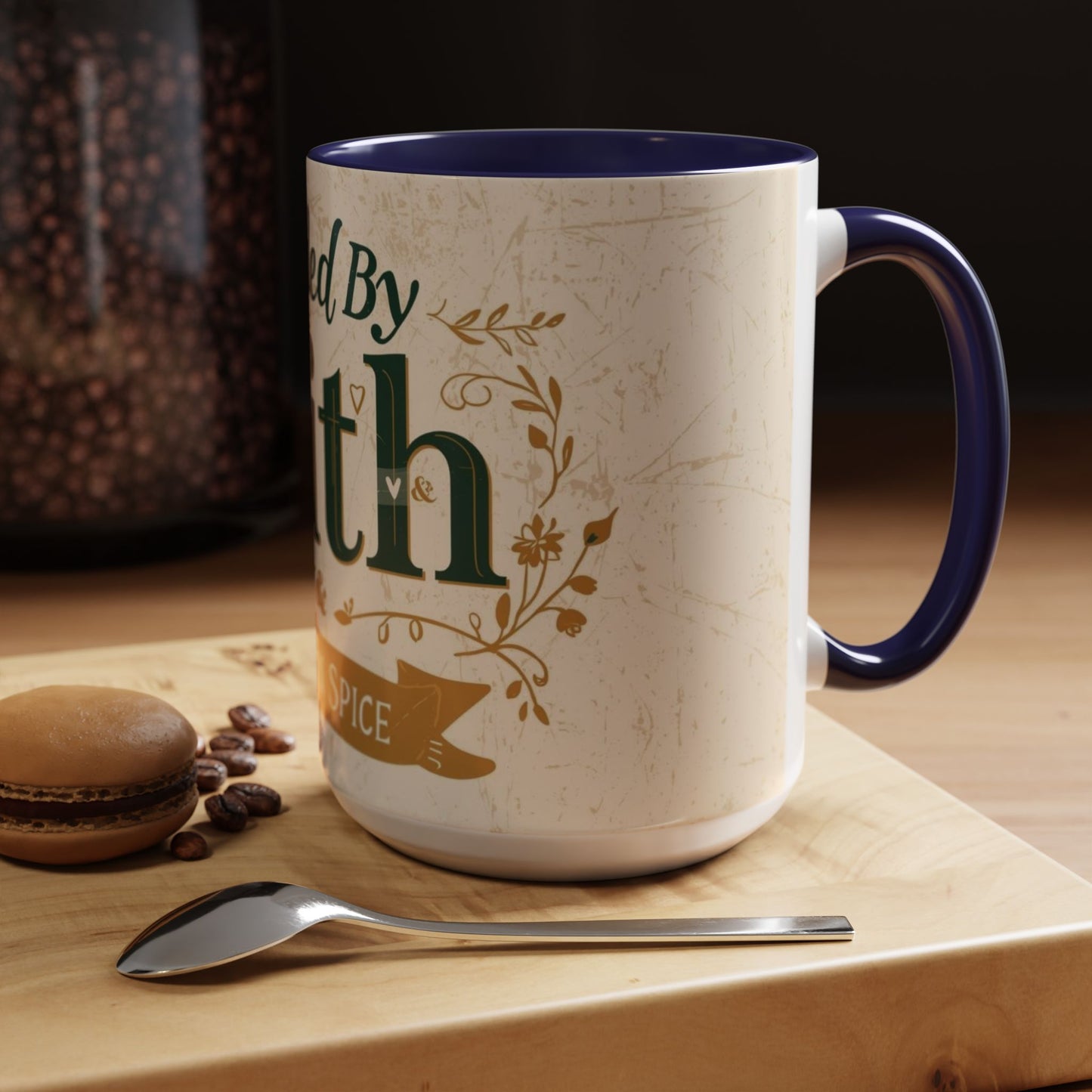 Lifestyle image of navy “Fueled by Faith & Pumpkin Spice” 15oz accent mug — Christian fall mug on wooden table beside macaron and coffee spoon.