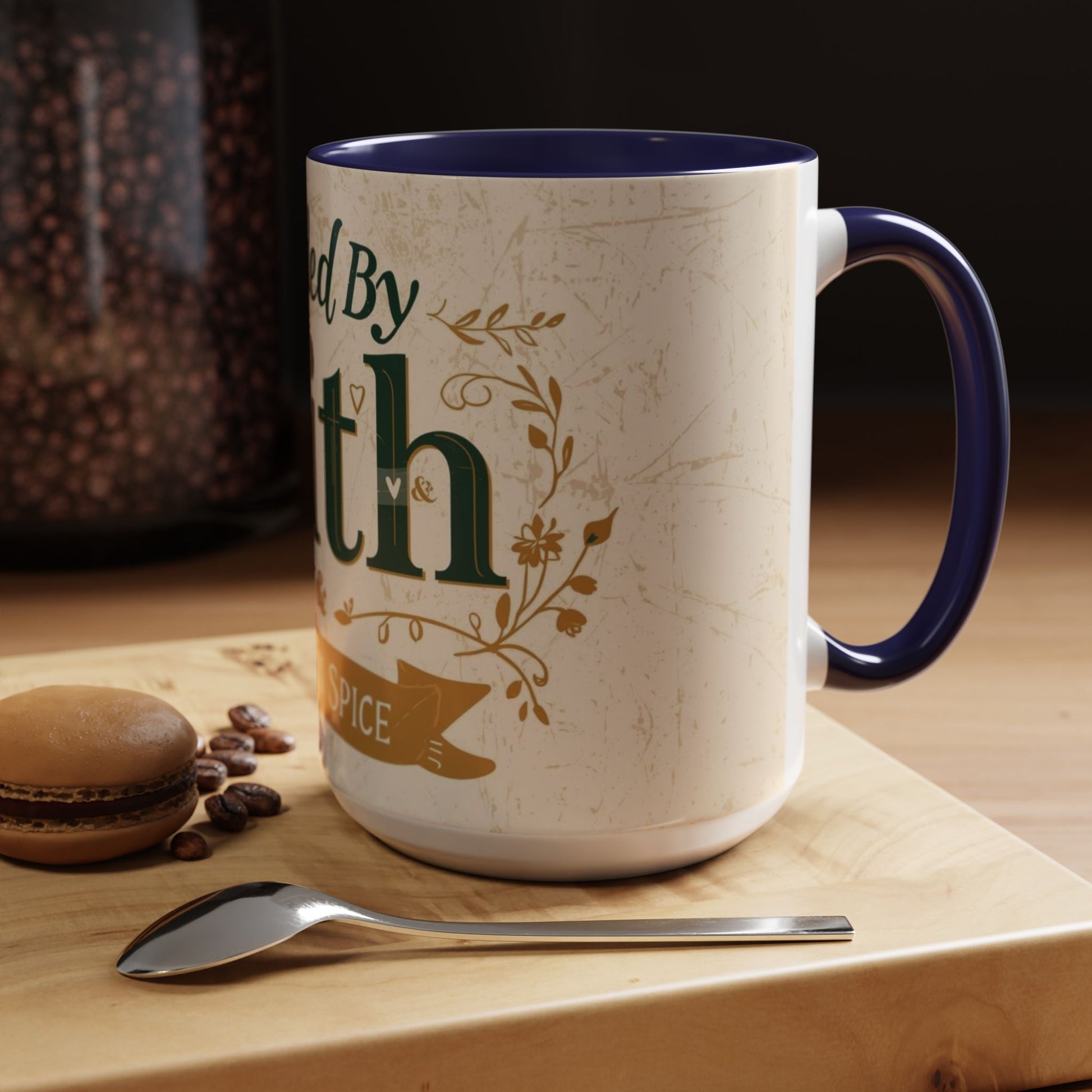 Lifestyle image of navy “Fueled by Faith & Pumpkin Spice” 15oz accent mug — Christian fall mug on wooden table beside macaron and coffee spoon.