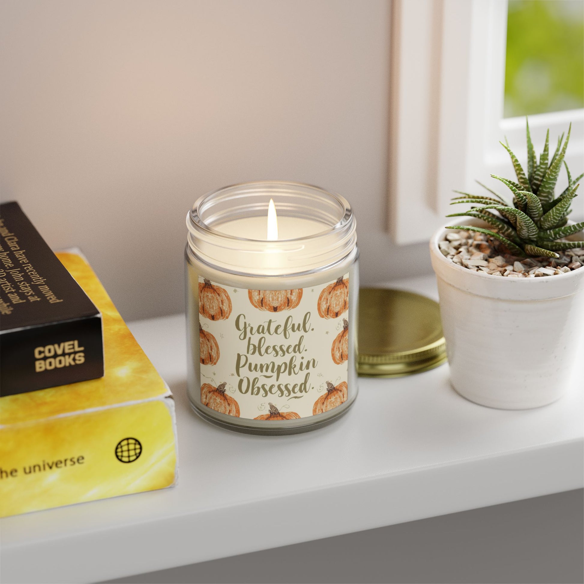 “Grateful, Blessed & Pumpkin Obsessed” clear jar candle glowing near books and plant — fall home accent candle with smooth beachwood aroma.