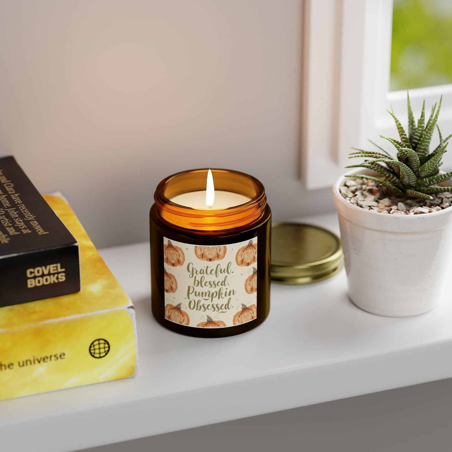 “Grateful, Blessed & Pumpkin Obsessed” amber candle glowing near books and plants — warm cinnamon chai candle for cozy fall home decor.