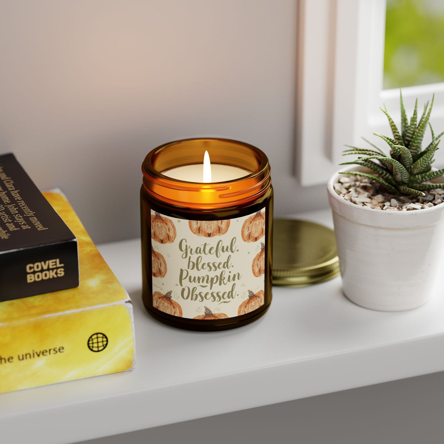 “Grateful, Blessed & Pumpkin Obsessed” amber jar candle glowing on shelf beside books and plant — warm fall candle with crisp Fraser Fir forest scent.